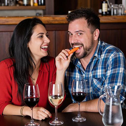 Photograph of a smiling couple at a wooden bar, woman in red blouse, man in blue plaid shirt, eating carrot, wine glasses in front