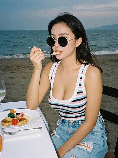 Photograph of an Asian woman with long black hair, wearing round sunglasses, white-striped tank top, and ripped jeans, eating at a beachside table