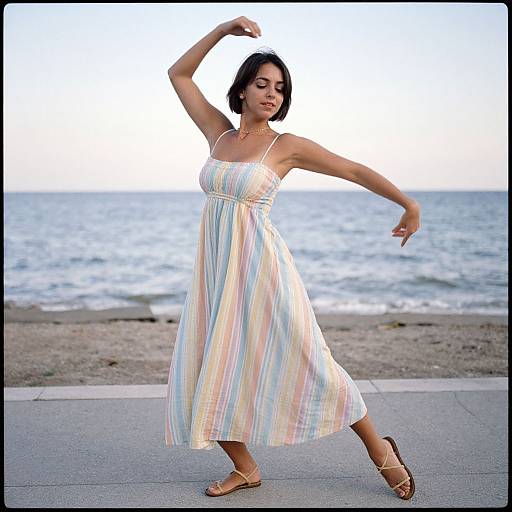 Photograph of a woman with short black hair, wearing a flowing, pastel-striped sundress, and sandals, dancing on a beachside sidewalk with
