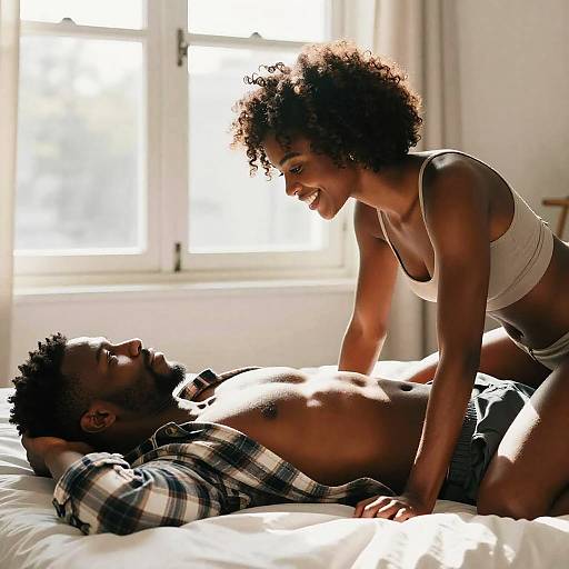 Photograph of a smiling Black couple in a sunlit bedroom; woman with curly hair in white bra and panties, man shirtless in plaid,