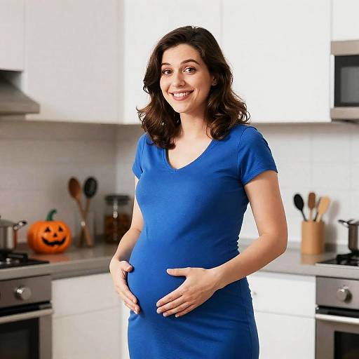 Pregnant woman with fair skin, brown wavy hair, wearing a blue dress, smiling in a modern white kitchen with a carved pumpkin on the