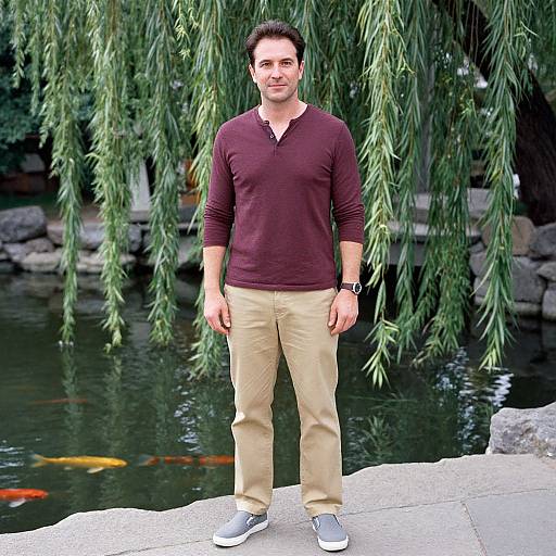 Photograph of a smiling man with short dark hair, wearing a maroon V-neck shirt, beige pants, and gray sneakers, standing by a tranquil