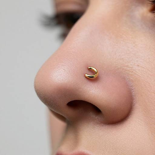 Close-up photograph of a light-skinned woman's nose with a small, gold, crescent-shaped nose stud. Background is blurred white.