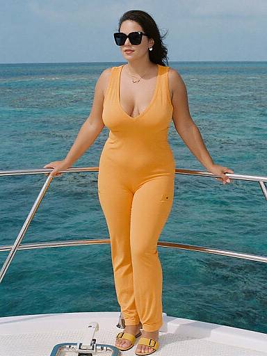 Photograph of a curvy woman with dark hair, wearing black sunglasses, a low-cut, bright orange jumpsuit, and yellow sandals, standing on