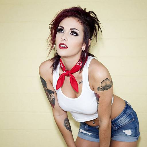 Photograph of a tattooed woman with dark hair, red lipstick, white tank top, red bandana, and denim shorts, leaning forward against a