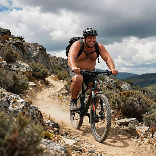 Muscular shirtless man in helmet and backpack mountain biking on rocky trail, surrounded by shrubs and mountains under cloudy sky.