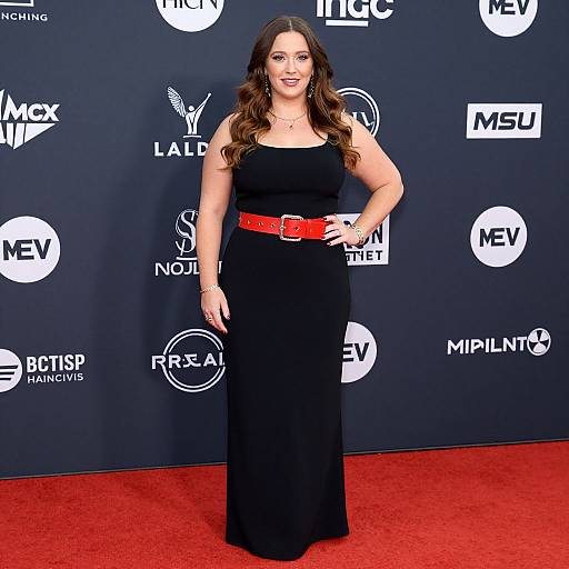 Photograph of a smiling woman with long brown hair, wearing a black sleeveless dress and red belt, standing on a red carpet against a black backdrop