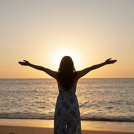 Silhouetted woman with arms outstretched, facing sunset over the ocean, wearing a long, patterned dress, standing on a sandy beach