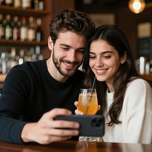 Photograph of a smiling couple with dark hair, the man in a black sweater and the woman in a white sweater, holding drinks while taking a selfie