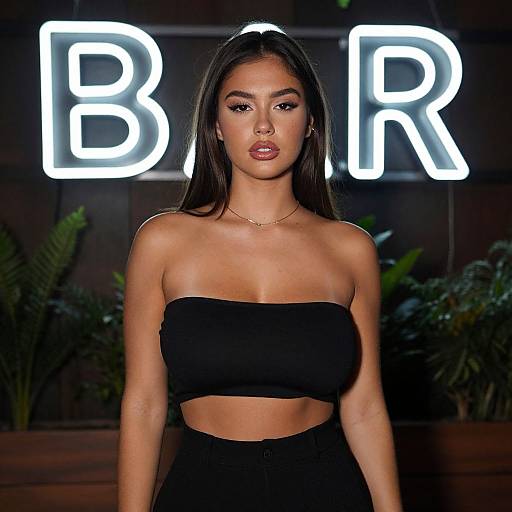 Photograph of a young woman with long dark hair, wearing a black strapless crop top and high-waisted pants, standing in front of a