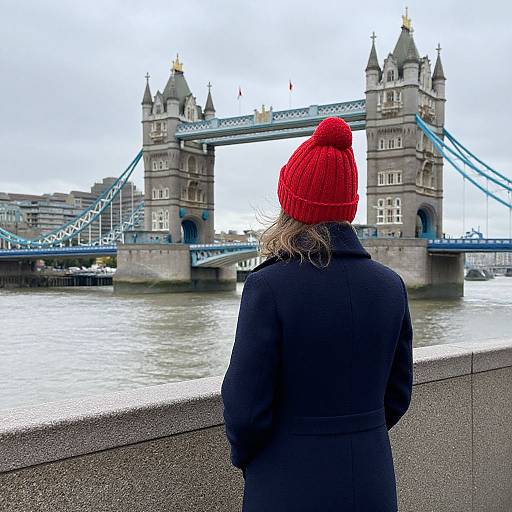 Photograph of a person in a red knit beanie and black coat, standing on a riverbank, facing London's Tower Bridge.