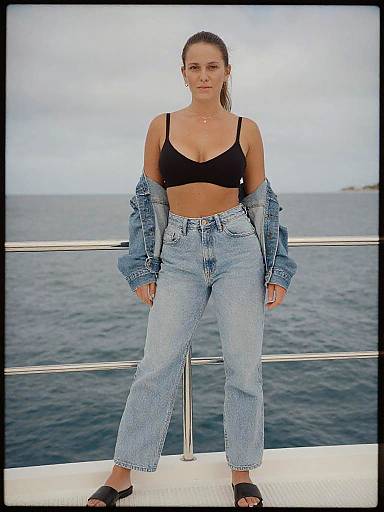 Photograph of a woman with a toned physique, wearing a black bra, blue high-waisted jeans, and black sandals, standing on a boat