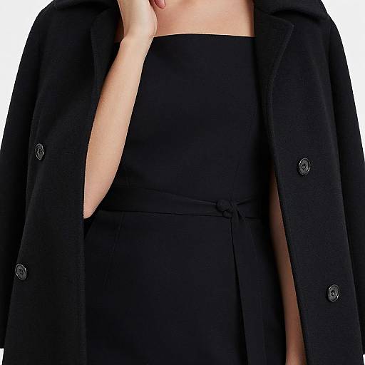 Photograph of a person wearing a black coat over a black dress, with one hand touching their chin, against a white background.