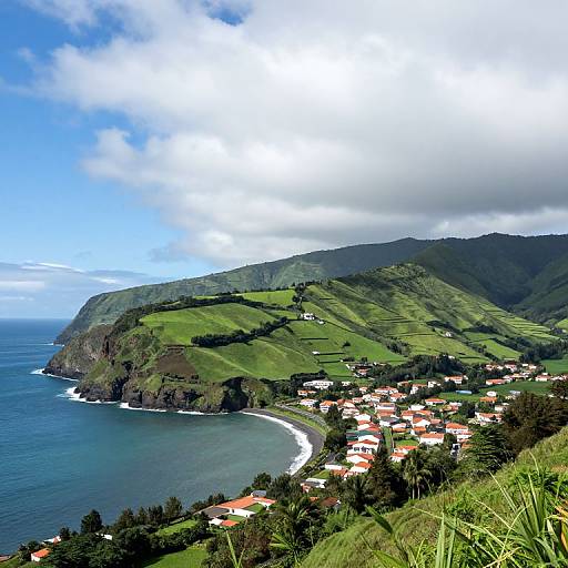 Photograph of a coastal hillside village with red-roofed houses, lush green fields, and a curved shoreline under a partly cloudy sky.