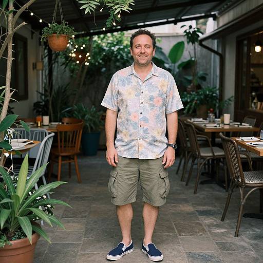 Photograph of a smiling, medium-build man in a floral shirt, khaki shorts, and navy slip-ons, standing in a lush, outdoor patio