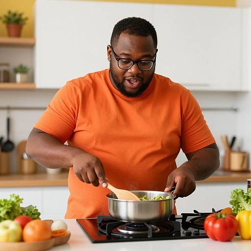 Photograph of a bearded, dark-skinned man in an orange shirt, wearing glasses, cooking in a bright kitchen with vegetables on the counter.