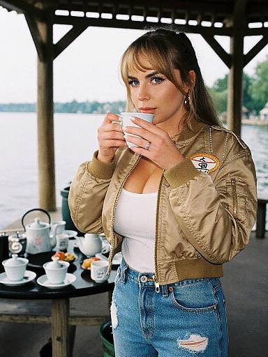 Photograph of a brunette woman with bangs, wearing a khaki bomber jacket, white tank top, and blue jeans, sipping tea under a