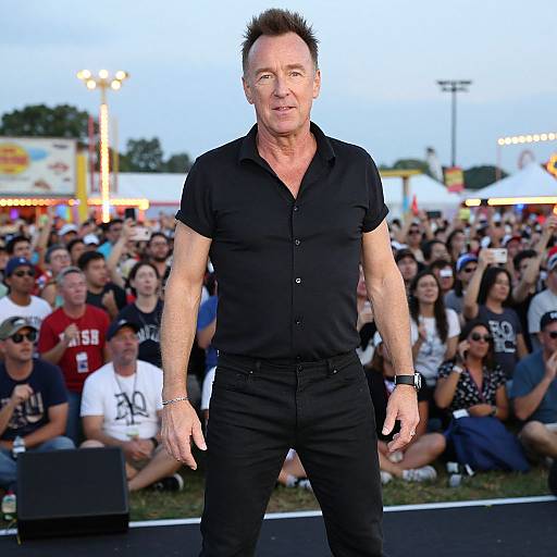 Photograph of a muscular, middle-aged man with spiky hair, wearing a black short-sleeve shirt and black pants, standing confidently in front