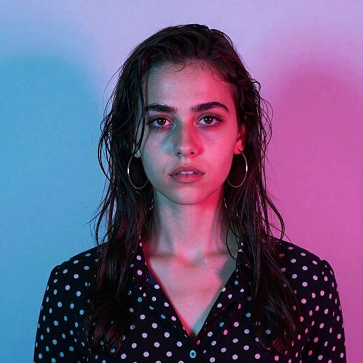 Photograph of a wet-haired woman with piercing gaze, wearing a black polka dot shirt, and large hoop earrings, illuminated by blue and pink neon