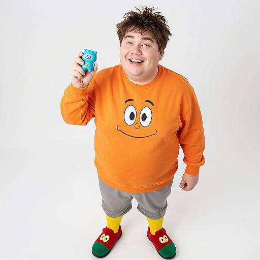 Photograph of a smiling, chubby man with spiky black hair, wearing an orange sweatshirt with a smiling face, gray pants, yellow socks,