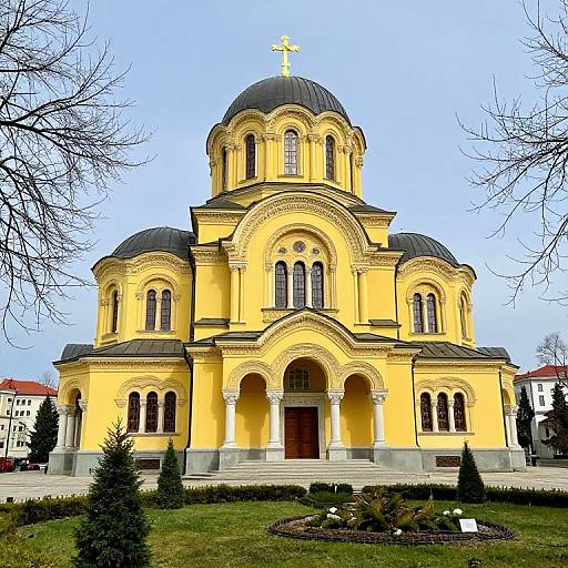 Photograph of a yellow Orthodox church with black domes and arched windows, set in a landscaped garden with bare trees.