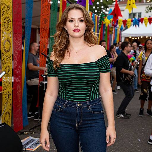 Photograph of a brunette woman with wavy hair, wearing an off-shoulder green striped top and dark jeans, standing in a colorful, festive