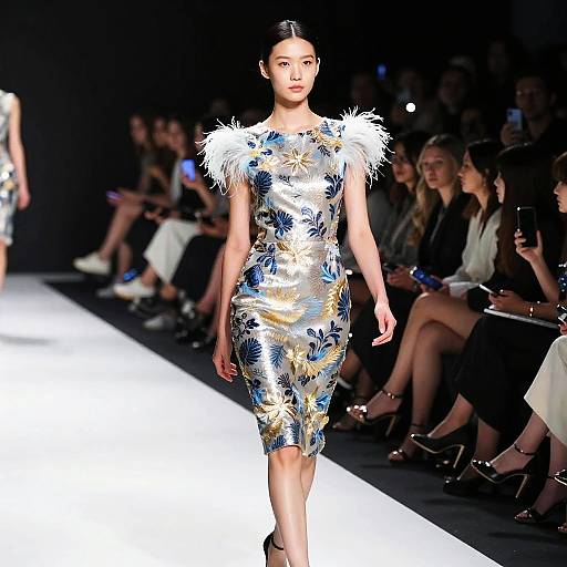 Photograph of an Asian woman with sleek black hair in a floral, metallic, form-fitting dress with white feather shoulders, walking a brightly lit runway