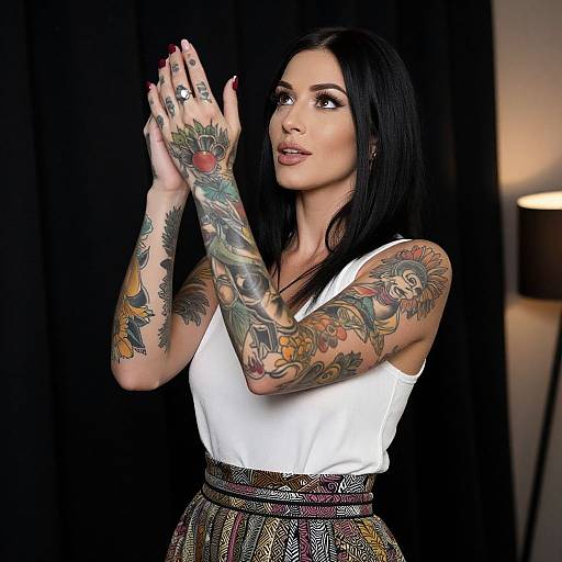 Photograph of a tattooed woman with dark hair, wearing a white sleeveless top and patterned skirt, hands raised in a praying pose, black