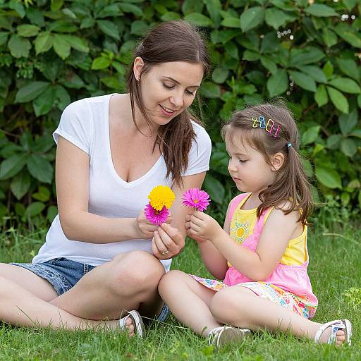 Photograph of a smiling woman with brown hair in a white shirt and denim shorts, sitting on grass, teaching a young girl with brown hair and a