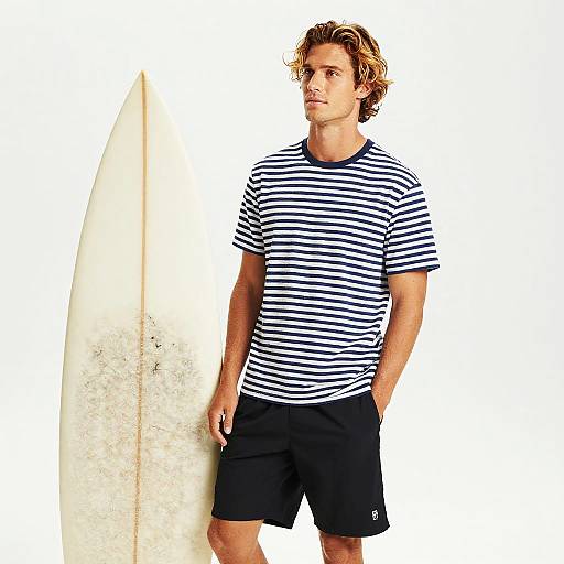 Photograph of a young, curly-haired man in a black-and-white striped shirt and black shorts, standing with a white surfboard.