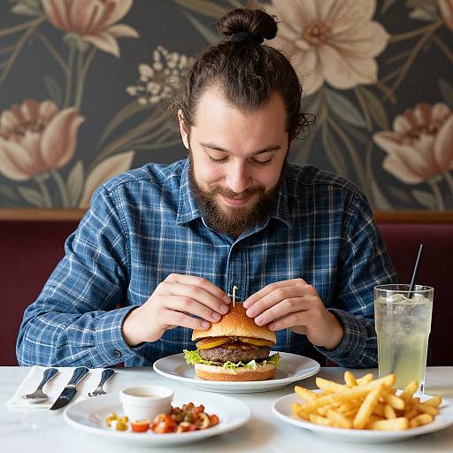 Photograph of a bearded man with a top knot, wearing a blue plaid shirt, assembling a burger with lettuce and cheese, surrounded by fries