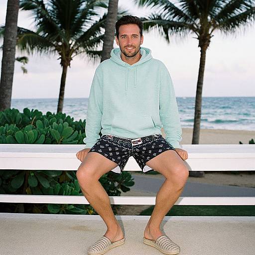 Photograph of a bearded man with short brown hair, wearing a light blue hoodie, black patterned shorts, and beige slip-on shoes, sitting