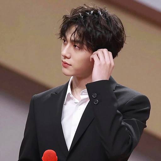 Photograph of a young Asian man with messy black hair, wearing a black suit over a white shirt, adjusting his hair while standing against a blurred indoor
