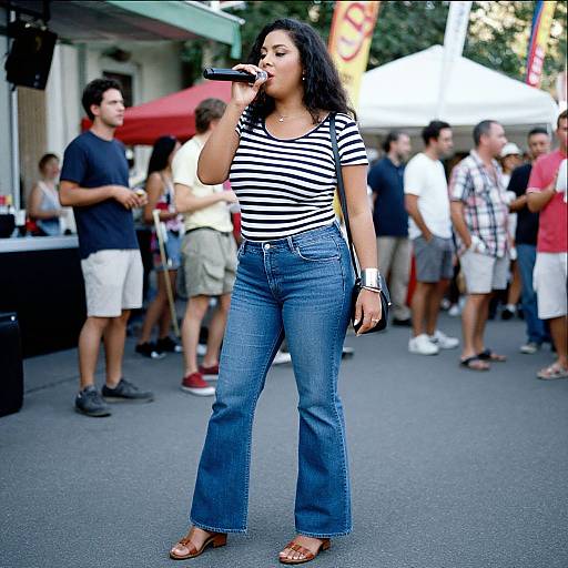 Photograph of a young woman with curly black hair, wearing a black-and-white striped shirt and blue jeans, singing into a microphone at an outdoor event