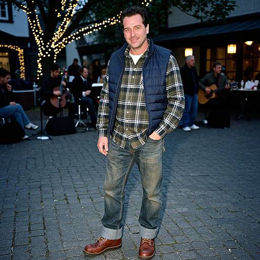 Photograph of a smiling man in a plaid shirt, blue vest, and denim jeans, standing on a cobblestone street at night with string