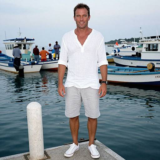 Photograph of a smiling, medium-build man in a white shirt and white shorts, standing on a dock with boats in the background. Wearing white