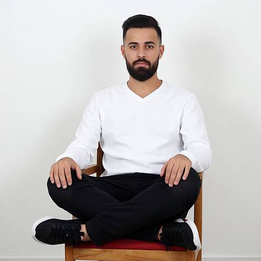 Photograph of a bearded man with short black hair, wearing a white long-sleeve shirt, black pants, and black shoes, sitting cross