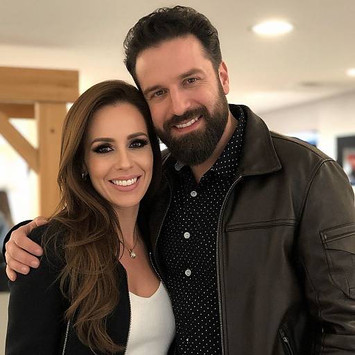 Photograph of a smiling, bearded man with dark hair and a black jacket, standing closely behind a woman with long brown hair, wearing a white