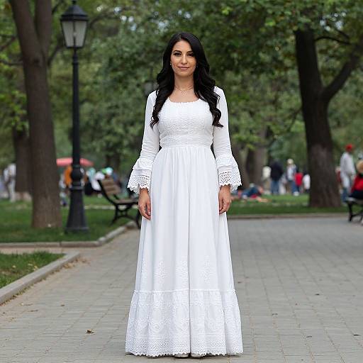 Photograph of a woman with long black hair, wearing a white, long-sleeve, lace-trimmed dress, standing on a park pathway