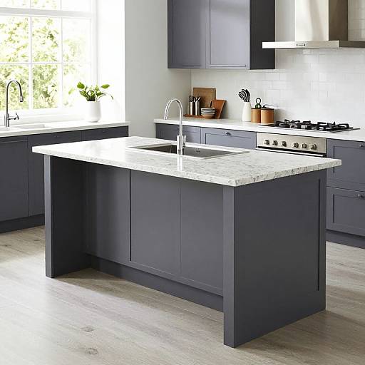 Photograph of a modern kitchen with dark blue cabinets, white marble island, stainless steel sink, and gas stove, under bright natural light.