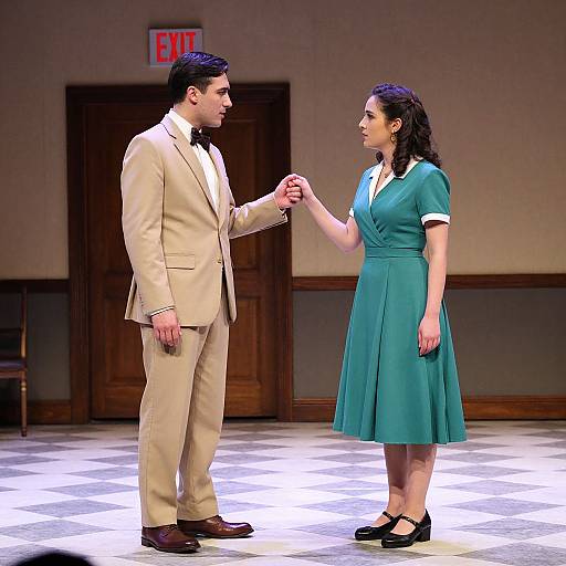 Photograph of a man in a beige suit and woman in a teal dress holding hands on a brightly lit stage, with an 