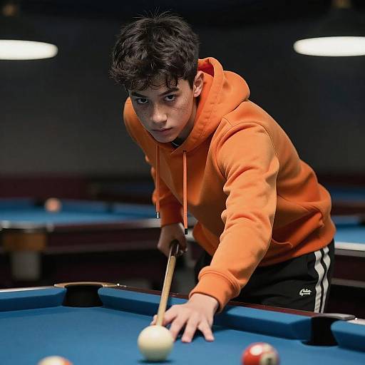 Photograph of a young boy with messy black hair, wearing an orange hoodie and black pants, aiming a pool cue at a ball on a blue pool