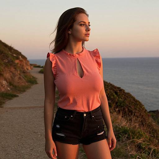 Photograph of a young woman with wavy brown hair, wearing a pink sleeveless top and black shorts, standing on a coastal path at sunset,