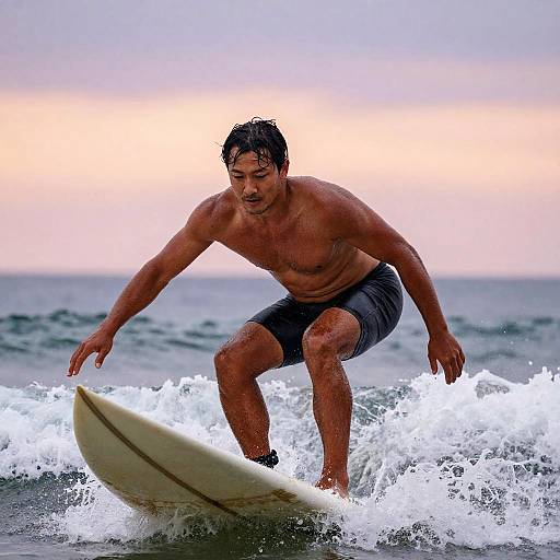 Photograph of a muscular, tan-skinned, shirtless man with wet black hair surfing on a white board in choppy ocean waves, wearing gray