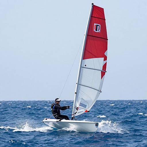 Photograph of a windsurfer in black wetsuit and white hat, sailing a red-and-white sail on blue ocean waves under clear sky.