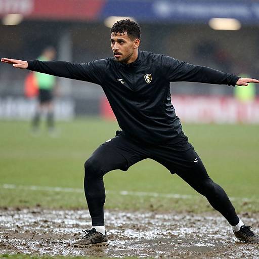 Photograph of a male soccer player with short curly hair, wearing a black tracksuit, stretching with arms wide on a muddy field. Blurred stadium