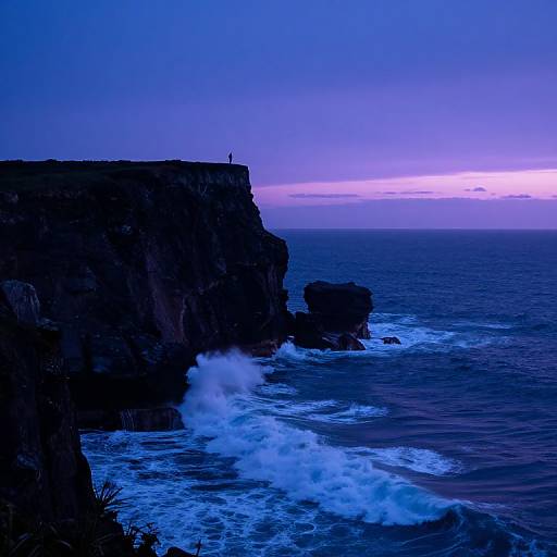 Photograph of a dark, rocky cliff at sunset, with a small figure standing on the edge, waves crashing below, and a vibrant blue-purple sky