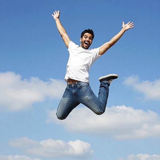 Photograph of a joyful man with dark hair, beard, white t-shirt, blue jeans, and black sneakers, jumping high against a bright blue sky