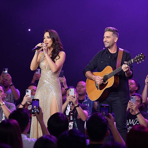 Photograph of a live performance: a woman in a shimmering gold dress sings into a microphone, while a man in black plays an acoustic guitar,