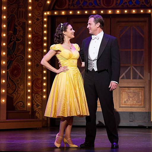Photograph of a couple on stage; woman in yellow dress, man in black tuxedo, smiling, illuminated theater backdrop.